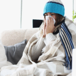 A woman sitting on a couch, wrapped in a blanket and wearing a hat, illustrating comfort during a cold.