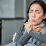 A woman sitting on a couch, mouth open, possibly expressing discomfort related to a sore throat.