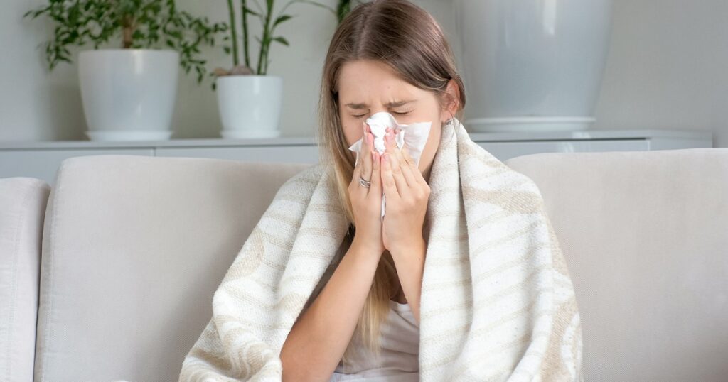 A young woman wrapped in a blanket sits on a couch, holding a tissue to her nose while sneezing, showing symptoms of flu or seasonal cold.