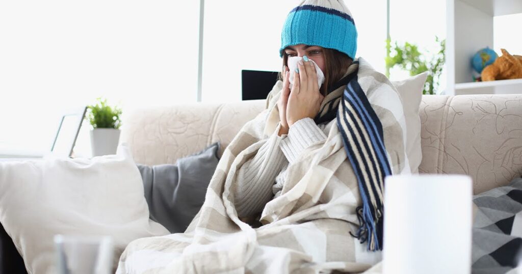 A woman sitting on a couch, wrapped in a blanket and wearing a hat, illustrating comfort during a cold.