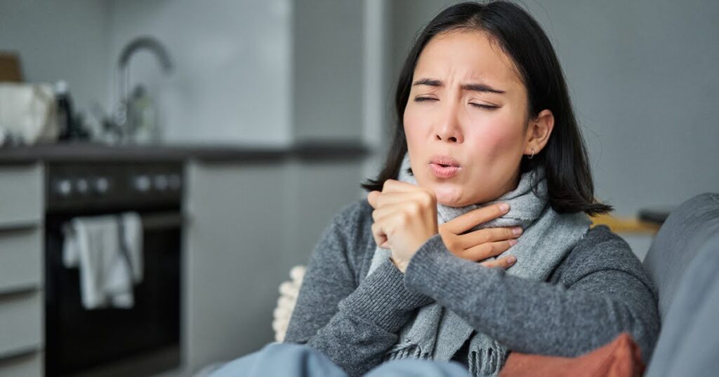 A woman sitting on a couch, mouth open, possibly expressing discomfort related to a sore throat.