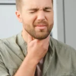 A close-up of a man with a beard squinting in pain while holding his hand to his throat, suggesting a sore throat or difficulty swallowing.
