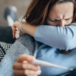 Woman with pneumonia coughing into her elbow while holding a thermometer.