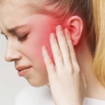 A young woman with blonde hair grimacing and holding her hand to her ear; a red glow is superimposed over the ear area to indicate an earache or inflammation.
