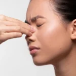 A woman with her hair in a ponytail pinching the bridge of her nose with her eyes closed, indicating sinus pressure, a headache, or a nosebleed.