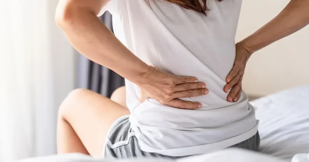 Close-up of a woman sitting on a bed holding her lower back in pain.