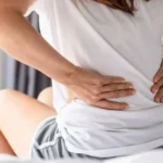 Close-up of a woman sitting on a bed holding her lower back in pain.
