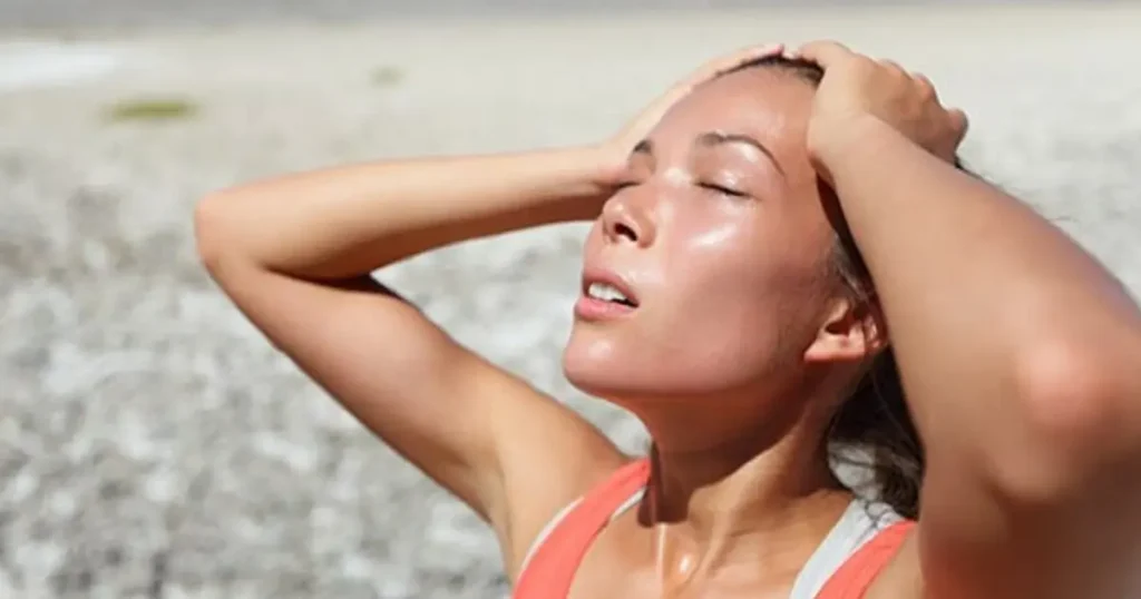A woman outdoors with her head tilted back and hands in her hair, looking exhausted and sweaty, suggesting heat exhaustion or a fever.