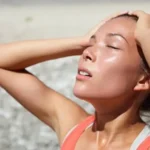 A woman outdoors with her head tilted back and hands in her hair, looking exhausted and sweaty, suggesting heat exhaustion or a fever.