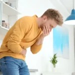 Man in a yellow sweater leaning against a kitchen counter holding his stomach.
