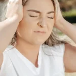 A woman in a white tank top outdoors holding both hands to the sides of her head with a pained facial expression, signifying a migraine or severe headache.