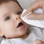 close-up of a baby lying down while an adult's hand gently wipes the baby's nose with a tissue, illustrating infant cold care.