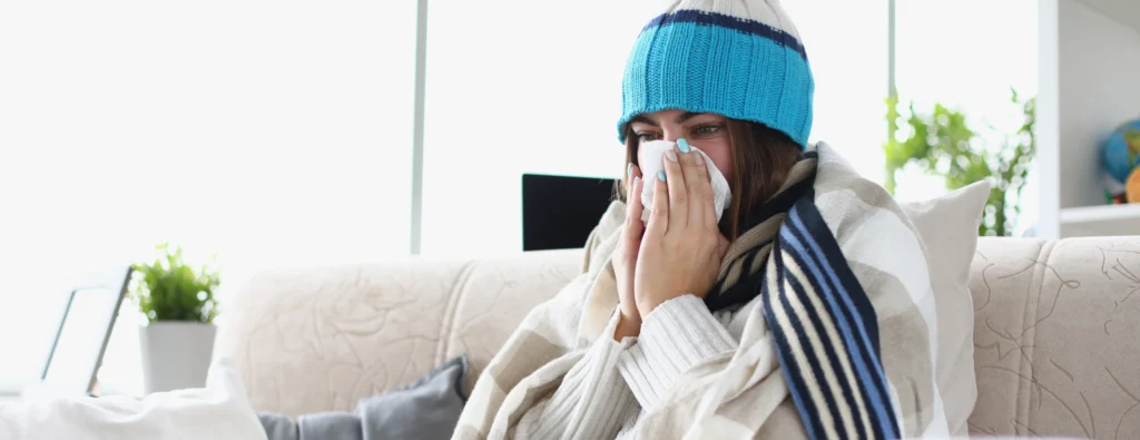 A woman sits on a couch wrapped in a blanket, wearing a blue beanie. She appears cold and uses a tissue, conveying an expression of discomfort.