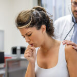Woman coughing during a medical exam while a doctor listens with a stethoscope, illustrating respiratory symptoms and clinical evaluation of a persistent cough.