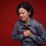 Woman in a grey jumpsuit clutching her chest with a pained expression on a red background, showing sudden chest pain or medical emergency.