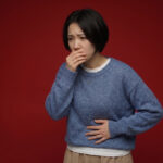 Woman in blue sweater with hand over mouth and stomach, showing nausea and discomfort on a dark red background.