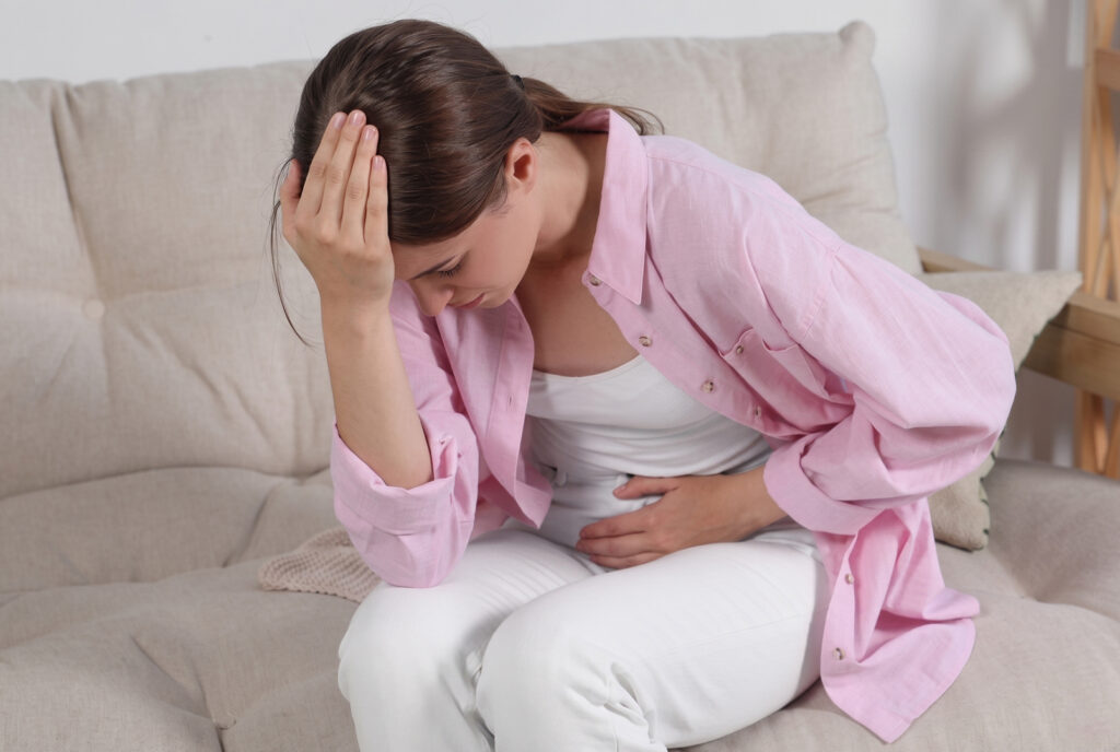 A woman sitting on a sofa while experiencing abdominal pain and a headache.