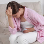 A woman sitting on a sofa while experiencing abdominal pain and a headache.