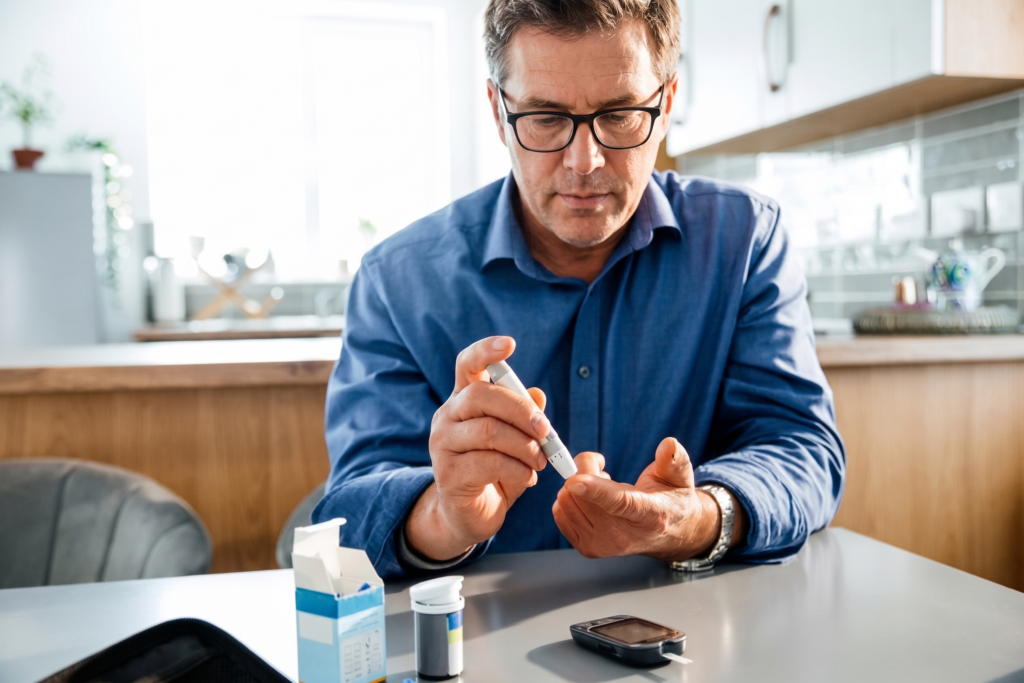 A middle-aged man sitting at a kitchen table testing his blood sugar with a lancing device and glucose meter.