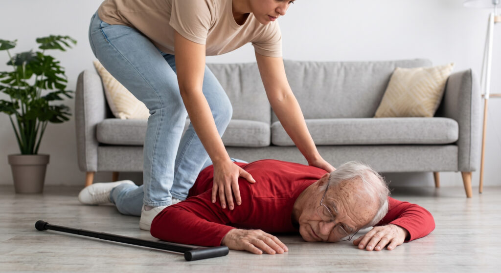 A younger person leans over to assist an elderly man who has fallen on a living room floor near his walking cane.