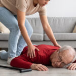 A younger person leans over to assist an elderly man who has fallen on a living room floor near his walking cane.