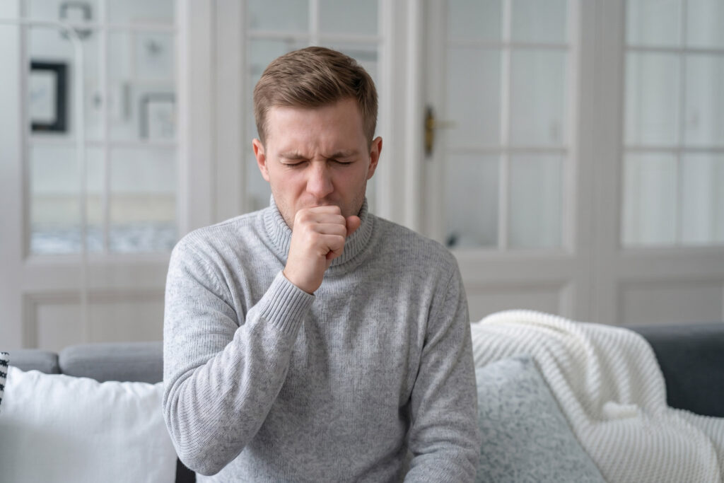 A close-up of a sick man showing symptoms of illness by coughing heavily into his hand, wincing with eyes closed.
