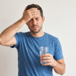 A man in a blue t-shirt holding his hand to his forehead while holding a glass of water.