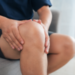 Realistic person holding swollen knee while sitting indoors with visible discomfort